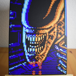 May include: A pixelated painting of the Alien from the film Alien, with a blue and black background. The Alien's head and mouth are visible, with sharp teeth. The painting is on a small wooden easel.