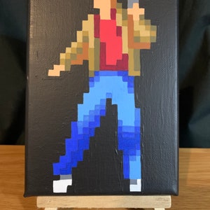 May include: Pixel art painting on canvas depicting a figure with a gun. The figure wears a red top, a brown jacket, and blue jeans. The background is black, and the painting is displayed on a small wooden easel.
