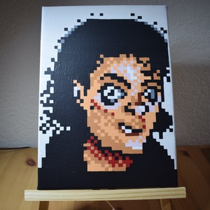 Könnte beinhalten: Ein Pixel-Art-Gemälde eines Gesichts auf einer kleinen Leinwandstaffelei. Die Person hat schwarze Haare, einen hellbraunen Teint und lächelt. Das Gemälde ist im Retro-8-Bit-Stil gehalten.