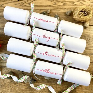 May include: White paper Christmas crackers with pink and green floral ribbon and personalised names in red script: Emma, Lucy, Steven, Catherine.