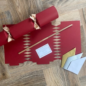 May include: A festive red Christmas cracker tied with gold ribbon, alongside a red paper template with a diamond pattern. A brown stick and small white card with text are also present. The text reads "Which of Santa's reindeer has the worst manners?"