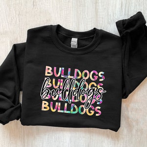 Op de afbeelding: Zwart sweatshirt met het woord "BULLDOGS" in een kleurrijk tie-dye ontwerp. Het woord "bulldogs" wordt drie keer herhaald, met een cursief lettertype in het midden. De sweatshirt heeft een ronde hals en lange mouwen.