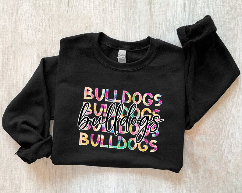 Op de afbeelding: Zwart sweatshirt met het woord "BULLDOGS" in een kleurrijk tie-dye ontwerp. Het woord "bulldogs" wordt drie keer herhaald, met een cursief lettertype in het midden. De sweatshirt heeft een ronde hals en lange mouwen.