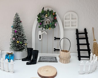 May include: A miniature fairy door with a wreath, a welcome mat, a small Christmas tree, a pair of black wellies, a small wooden bucket, a black ladder, and a white picket fence.