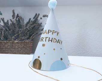 Op de afbeelding: Witte papieren feestmuts met een blauwe pompon bovenop. De muts heeft gouden letters met de tekst "Happy Birthday" en blauwe en gouden sterren.