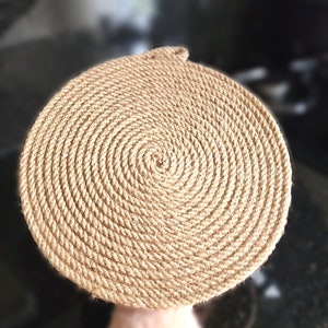 May include: A round, natural fibre coaster with a spiral pattern. The coaster is made of woven rope and has a light brown colour.