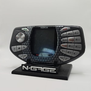 May include: A black Nokia N-Gage handheld gaming console and mobile phone featuring a central screen, a directional pad on the left, and a numeric keypad on the right. The device is displayed on a black stand with the N-Gage logo in white text.