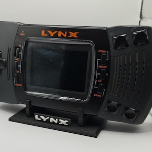 May include: A black handheld video game console with orange LYNX branding above the screen. The device features a directional pad, A and B buttons, and various control buttons labeled Power, Backlight, Option 1, Restart, Flip, and Option 2.