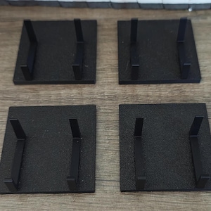 May include: Four black square display stands, each featuring a flat base and two vertical L-shaped supports. These matte black holders are designed to hold items upright, providing a stable foundation for various objects in a set of four pieces.