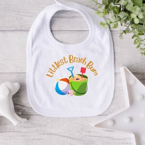 May include: White baby bib with the text "Littlest Beach Bum" in orange script. The bib features a colorful beach scene with a bucket, shovel, ball, and seashells. A perfect accessory for a day at the beach.
