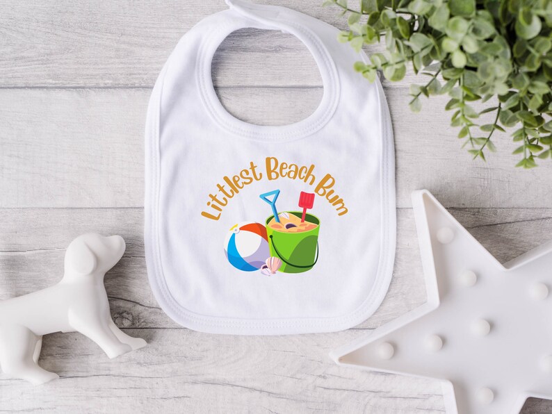 May include: White baby bib with the text "Littlest Beach Bum" in orange script. The bib features a colorful beach scene with a bucket, shovel, ball, and seashells. A perfect accessory for a day at the beach.