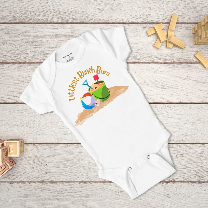Littlest Beach Bum, featuring a beach ball and bucket of sand, sitting on the sand.  Shown on a white onesie.