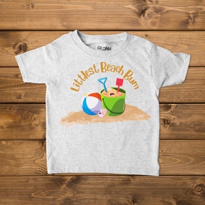 Littlest Beach Bum, featuring a beach ball and bucket of sand, sitting on the sand. Shown on a child's t-shirt.