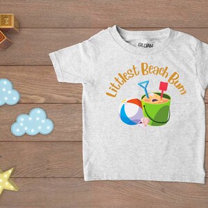 Littlest Beach Bum, featuring a beach ball and bucket of sand.  Shown on a white t-shirt.