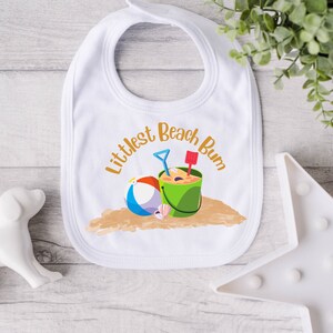 Littlest Beach Bum, featuring a beach ball and bucket of sand, sitting on the sand.  Shown on a white baby bib.