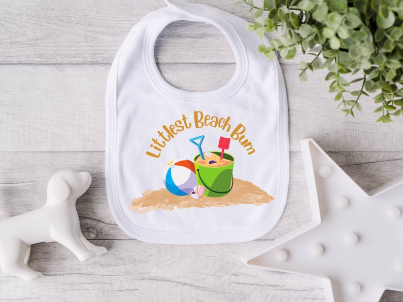 Littlest Beach Bum, featuring a beach ball and bucket of sand, sitting on the sand.  Shown on a white baby bib.