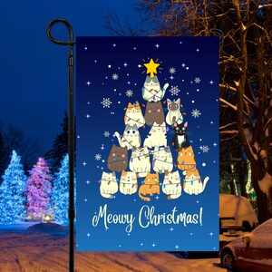 May include: A blue Christmas-themed garden flag with a Christmas tree made of cats. The cats are wearing Christmas lights and are stacked on top of each other. The text "Meowy Christmas!" is at the bottom of the flag.