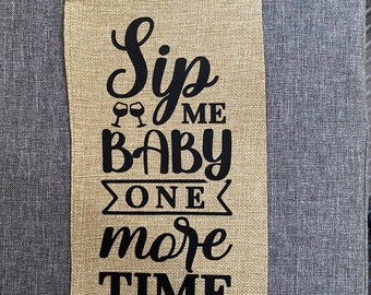 以下が含まれることがあります： 「Sip Me Baby One More Time」という黒い文字が書かれたジュート製のワインボトルバッグです。