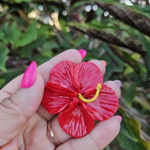 May include: A vibrant red hibiscus flower with a yellow stamen is held in a hand. The flower has detailed petals and a glossy finish. The background is blurred green foliage.