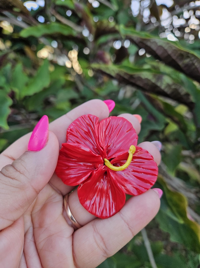 May include: A vibrant red hibiscus flower with a yellow stamen is held in a hand. The flower has detailed petals and a glossy finish. The background is blurred green foliage.