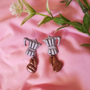May include: A pair of silver coffee pot earrings with brown coffee bean charms. The earrings are on a pink fabric background.