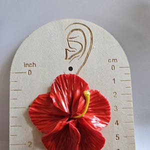 May include: A vibrant red hibiscus flower earring, with a yellow stamen, is displayed on a wooden earring gauge. The gauge shows measurements in inches and centimeters. The earring is approximately 2 inches in diameter.