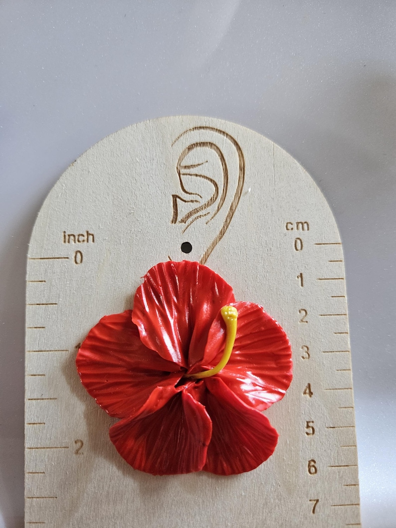 May include: A vibrant red hibiscus flower earring, with a yellow stamen, is displayed on a wooden earring gauge. The gauge shows measurements in inches and centimeters. The earring is approximately 2 inches in diameter.