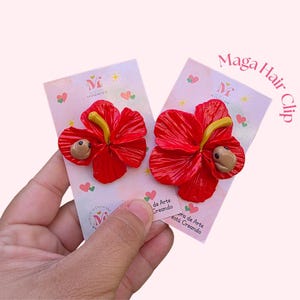 May include: Two red hibiscus flower hair clips with yellow stamens and a small brown creature accent. The clips are displayed on pink cards with hearts and the text "Maga Hair Clip". The cards also have the text "Maga Handmade".
