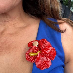 May include: A red flower brooch with a yellow stamen and a small, light-colored figure in the center. The brooch is pinned to a blue garment.