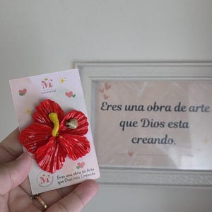 May include: A handmade red hibiscus flower brooch with a yellow stamen and a small insect detail. The brooch is attached to a card with the text "Eres una obra de Arte que Dios est&aacute; Creando."