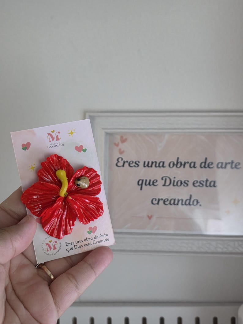 May include: A handmade red hibiscus flower brooch with a yellow stamen and a small insect detail. The brooch is attached to a card with the text "Eres una obra de Arte que Dios est&aacute; Creando."