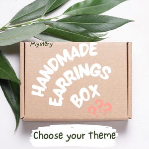 Puede incluir: Una caja de cartón marrón con las palabras "HANDMADE EARRINGS BOX" en blanco. La caja también tiene la palabra "Mystery" y los signos de interrogación "??" en rosa. La frase "Choose your theme" está escrita debajo.