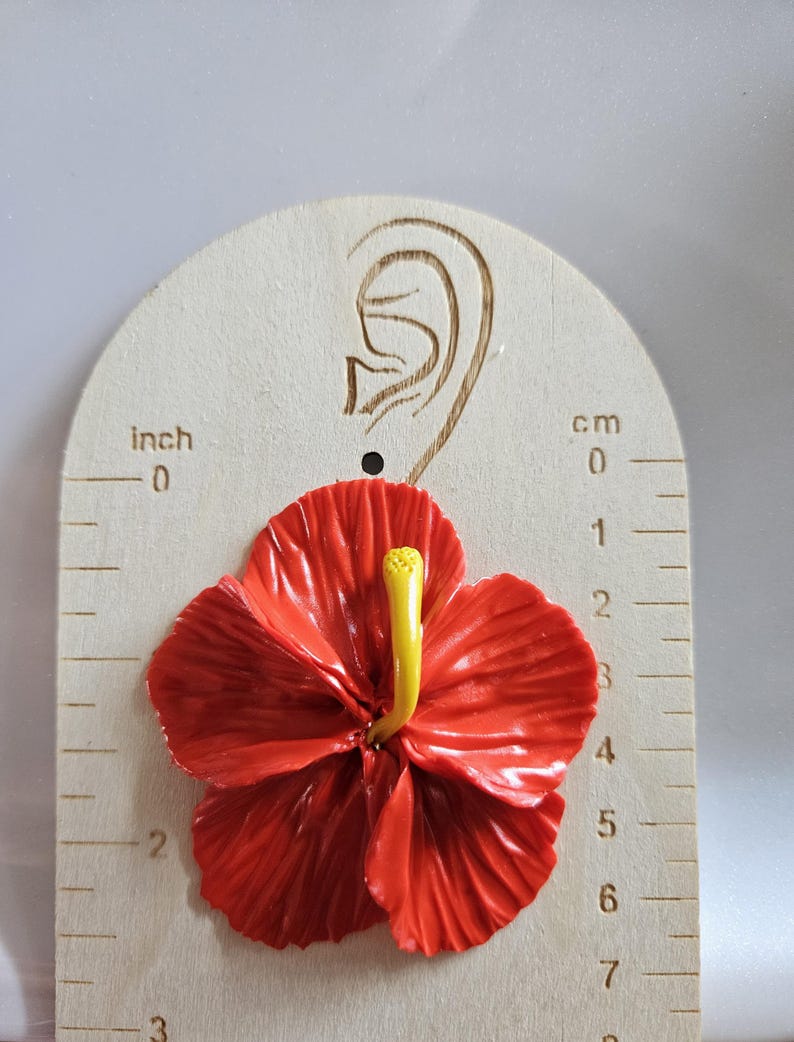 May include: A vibrant red hibiscus flower, with a yellow stamen, is displayed on a wooden measuring tool. The tool has inch and centimeter markings. The flower is approximately 2 inches in diameter.