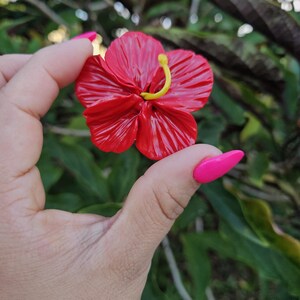 May include: A vibrant red hibiscus flower with a yellow stamen. The flower has detailed petals and is held against a blurred green background. The flower is a bright, eye-catching piece.