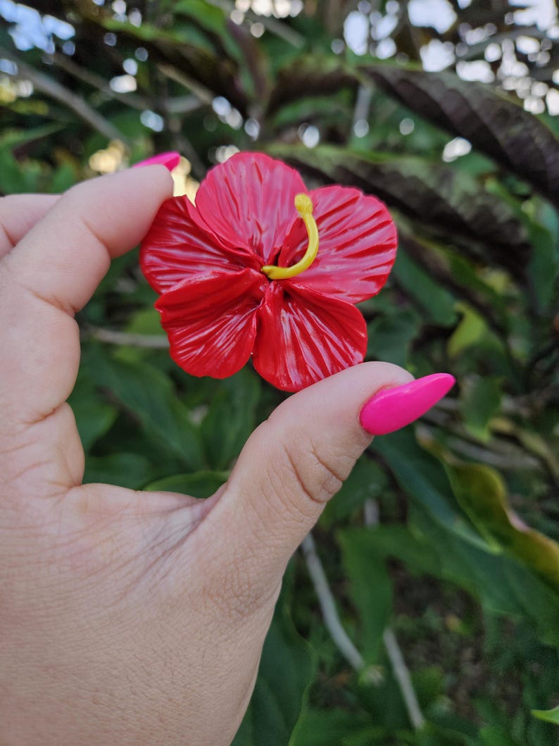 May include: A vibrant red hibiscus flower with a yellow stamen. The flower has detailed petals and is held against a blurred green background. The flower is a bright, eye-catching piece.