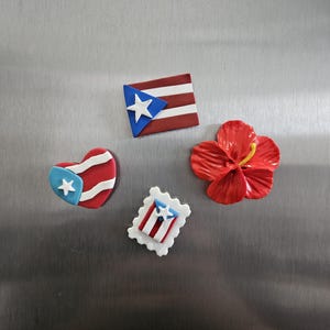 May include: A collection of colorful magnets featuring Puerto Rican flag designs and a red hibiscus flower. The magnets include a heart, a rectangular flag, and a postage stamp design, all with the flag's colors and symbols.