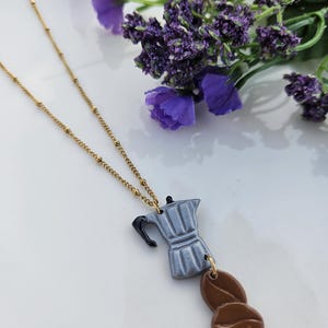 May include: A gold chain necklace with a silver-colored ceramic coffee pot charm and a brown ceramic coffee bean charm.