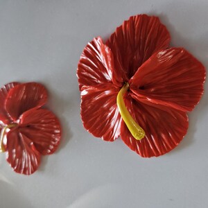 May include: Two red hibiscus flowers with yellow stamens are displayed against a light gray background. The larger flower is in full bloom, while the smaller one is partially open. The petals have a textured appearance.