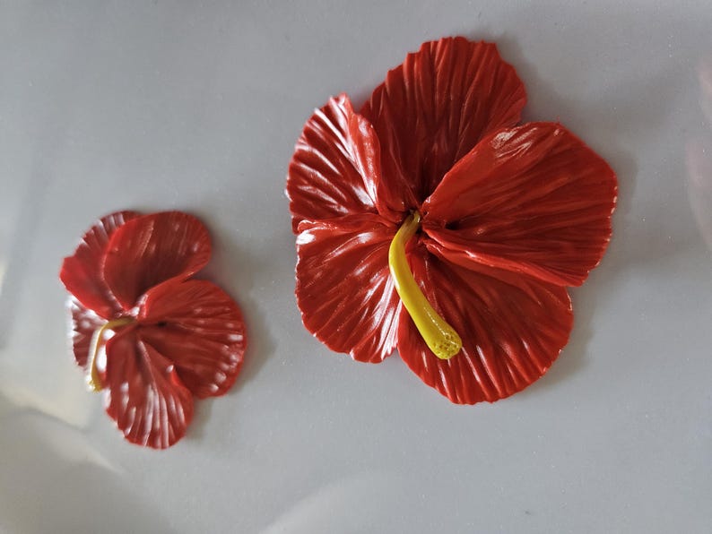 May include: Two red hibiscus flowers with yellow stamens are displayed against a light gray background. The larger flower is in full bloom, while the smaller one is partially open. The petals have a textured appearance.