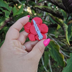 May include: A vibrant red flower hair clip with a silver metal barrette. The flower has multiple petals and is held in a hand with pink nail polish. The background is blurred green foliage.