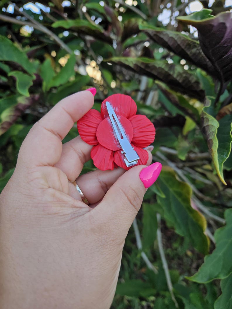 May include: A vibrant red flower hair clip with a silver metal barrette. The flower has multiple petals and is held in a hand with pink nail polish. The background is blurred green foliage.