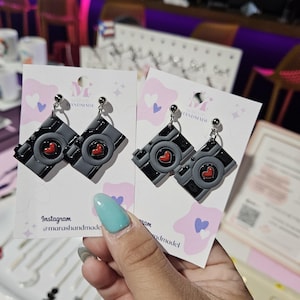 May include: Two pairs of black camera-shaped earrings with red hearts on the front. The earrings are on white cards with pink hearts and the text "Mi Handmade" on them. The cards also have the text "Instagram @marashandmade" on them.