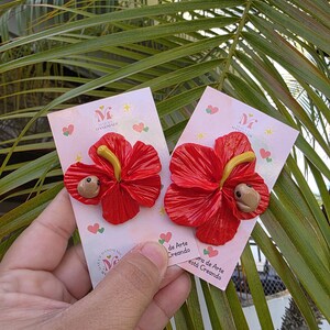 May include: Two handmade red hibiscus flower earrings with yellow stamens and a small brown creature. The earrings are displayed on pink cards with the text "Handmade" and "de Arte Creando". The background features green palm leaves.