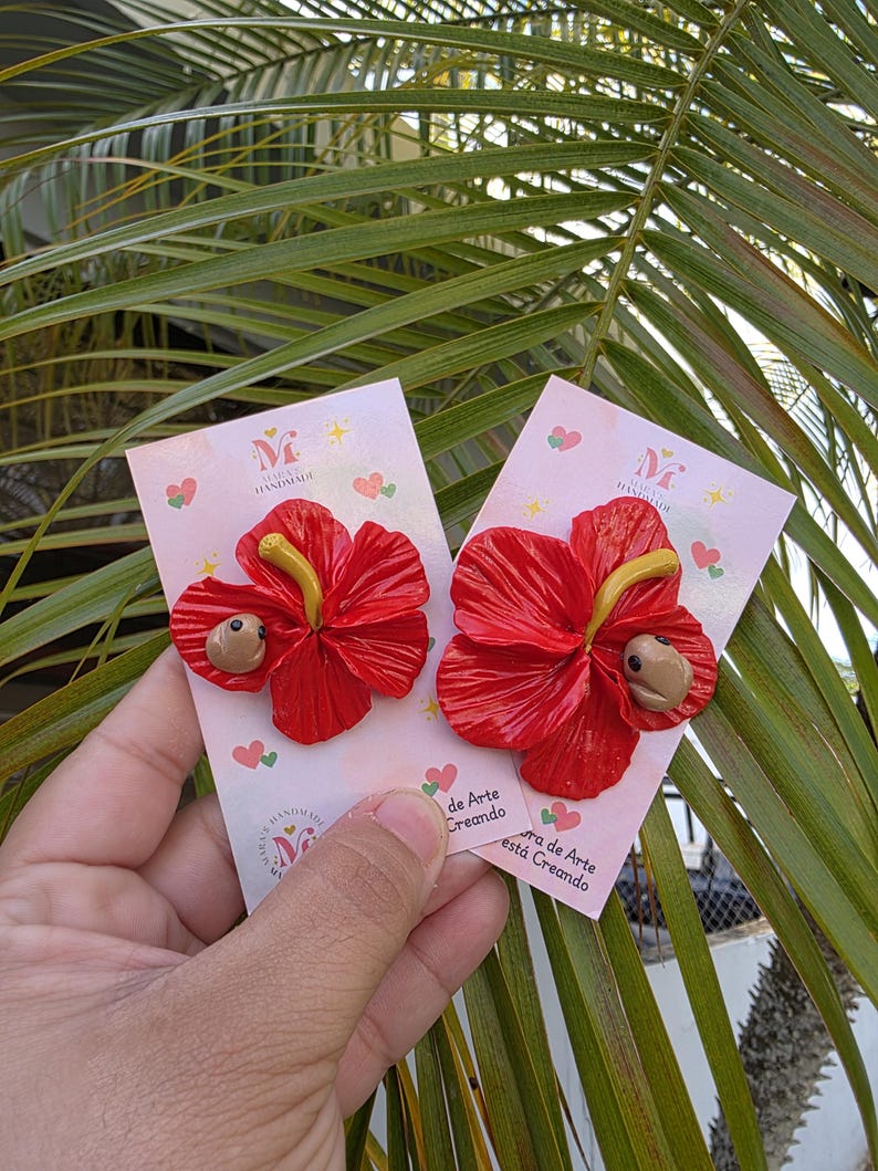 May include: Two handmade red hibiscus flower earrings with yellow stamens and a small brown creature. The earrings are displayed on pink cards with the text "Handmade" and "de Arte Creando". The background features green palm leaves.