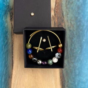 May include: A set of gold-colored hoop earrings and two matching rings with a small gemstone in a black box. The earrings are adorned with colorful beads. A small gemstone stud is also in the box.