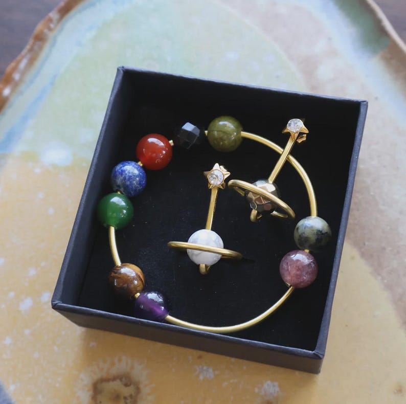 May include: A black box contains a set of celestial-themed jewelry. The set includes a ring with a white orb, a pair of earrings with star-shaped accents, and a bracelet with colorful gemstone beads. The jewelry has gold-colored metal.