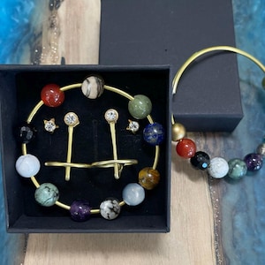 May include: A jewelry set featuring a gold-toned bracelet and earrings. The bracelet and the earrings' frames are adorned with colorful, spherical beads. The earrings include a pair of star-shaped studs and a pair of long, thin earrings with a single gem.