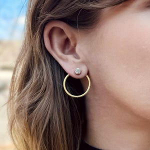 May include: A close-up of a person's ear adorned with two gold earrings. One is a small stud with a clear stone, and the other is a large, textured gold hoop earring. The hoop earring is approximately 1.5 inches in diameter.