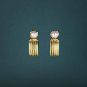 May include: A pair of gold-toned earrings featuring a smooth, round white pearl at the top. Below each pearl, the earring design consists of three parallel, curved gold-toned metal bars that form a semi-circular, open-ended loop structure.