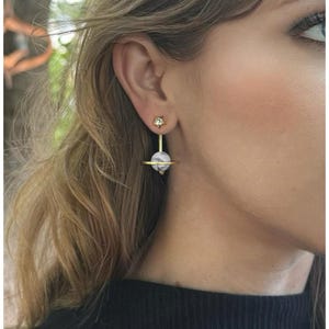 May include: Gold-tone earrings with a celestial design. Each earring features a small star-shaped stud, a vertical gold bar, and a round, marbled stone. A gold ring encircles the stone. The earrings are a unique accessory.